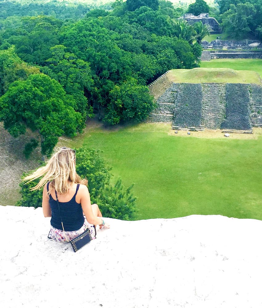 A person sits on a ledge overlooking a lush green landscape and ancient ruins.