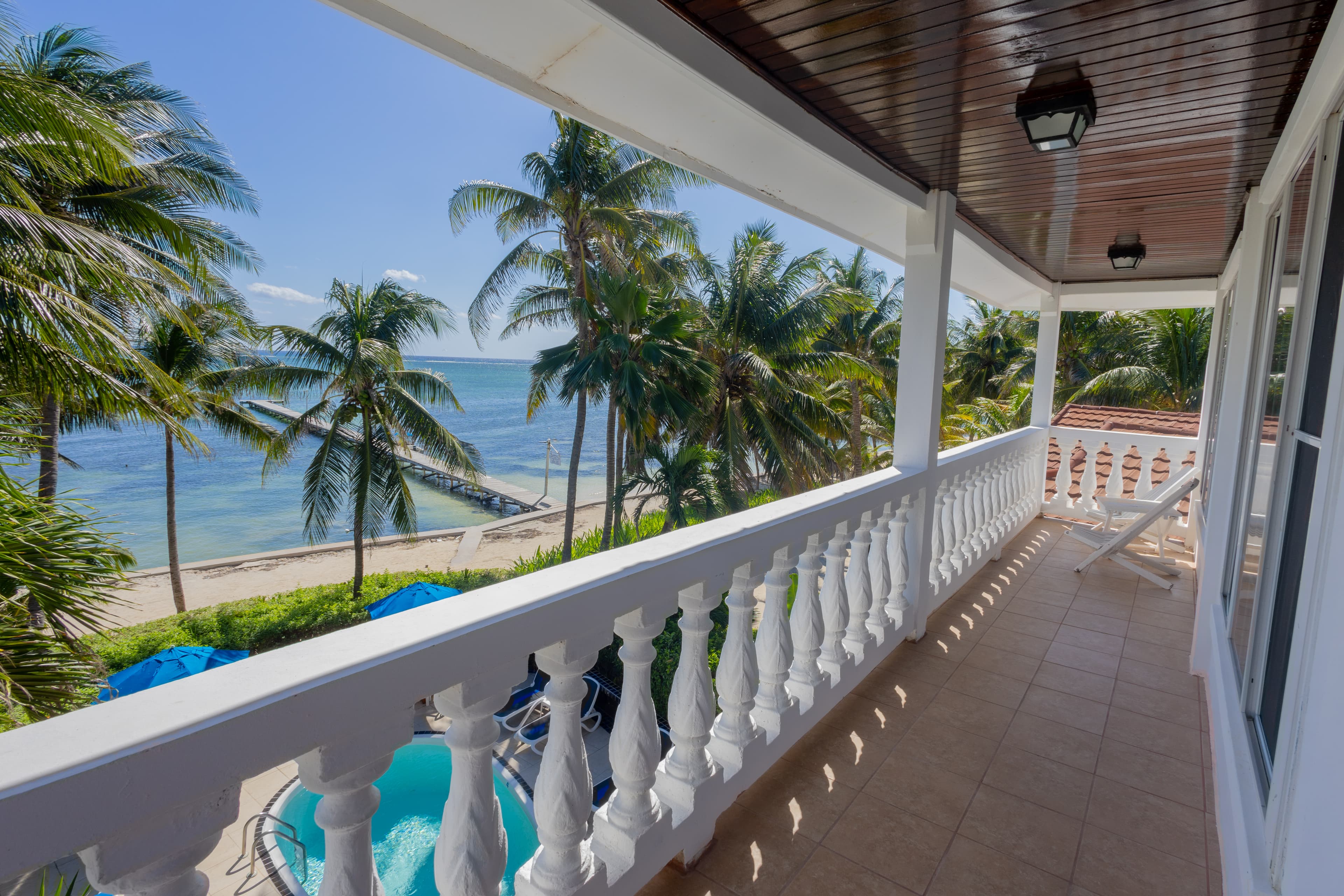 A sunlit balcony overlooking a beach with palm trees and a blue ocean.
