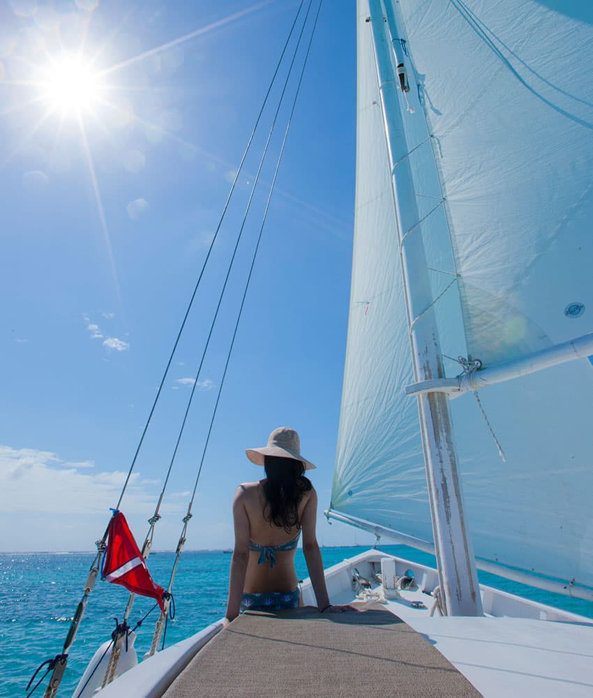 A person sits on the bow of a sailboat under a bright sun, overlooking the turquoise water.