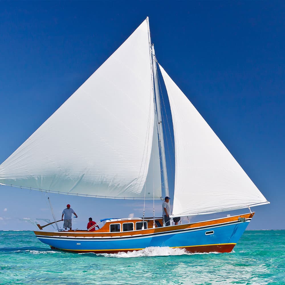 A sailboat with three people navigates across clear blue waters under a bright sky.