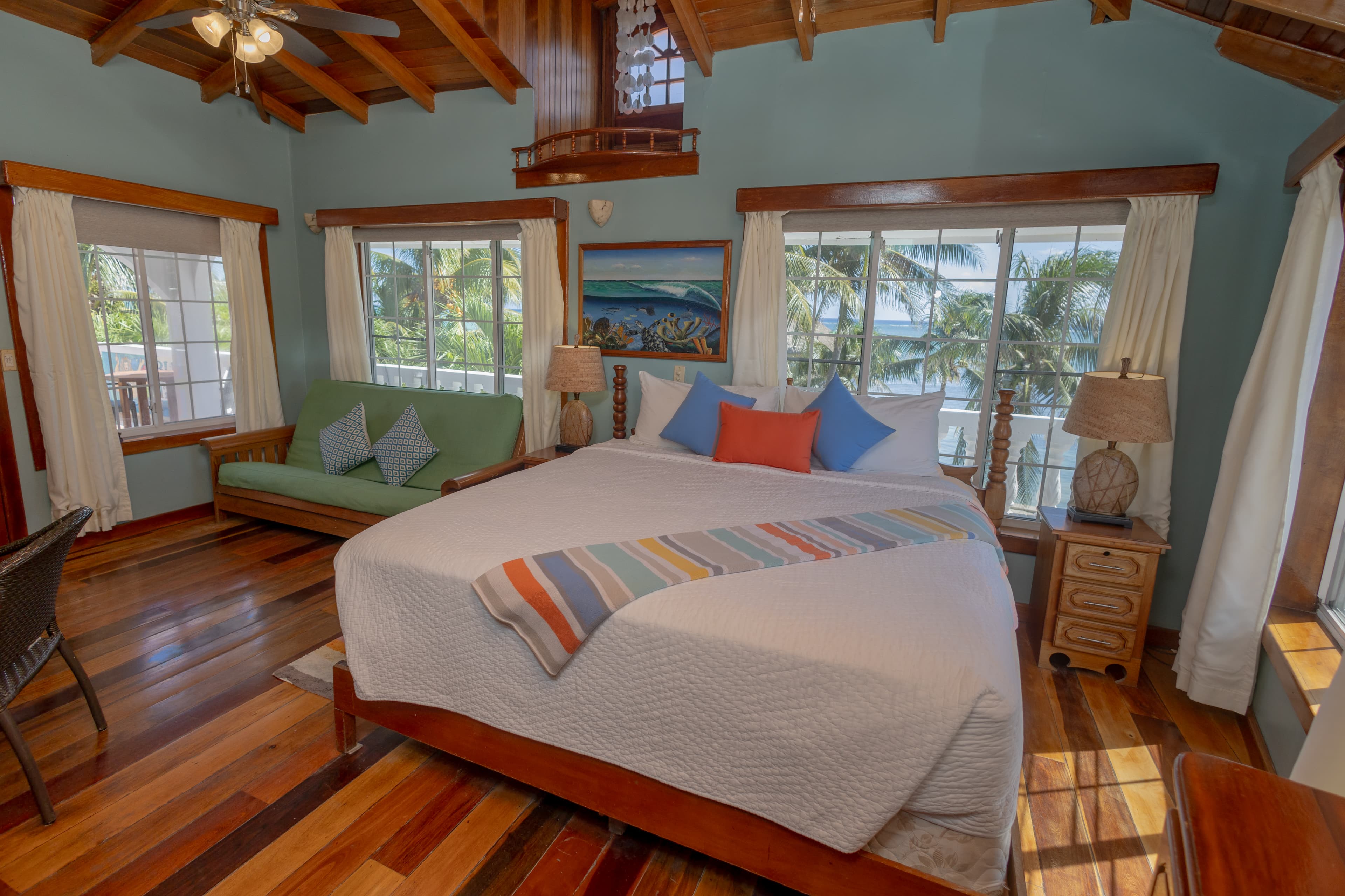 Cozy bedroom with a queen bed, vibrant pillows, a green couch, and large windows overlooking palm trees.